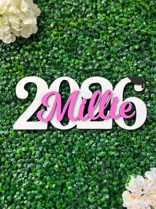 Decorative sign with '2026 Millie' on a green textured background with white flowers.