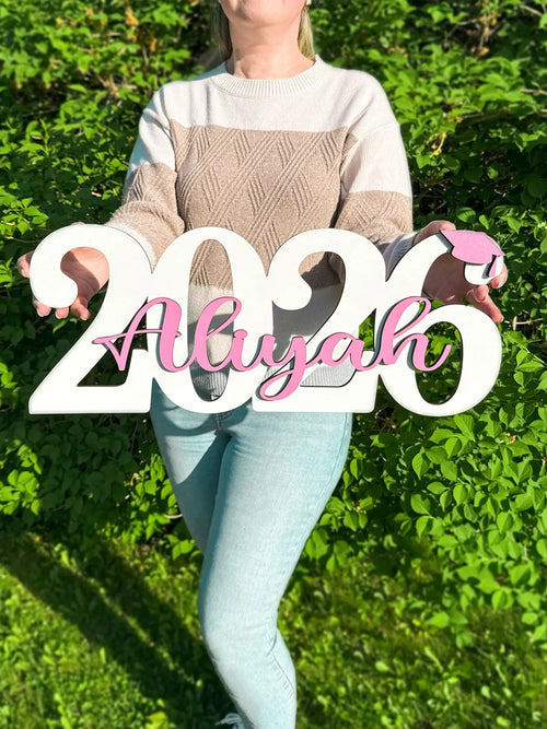 Person holding a decorative sign with '2026 Aliyah' against a green outdoor background