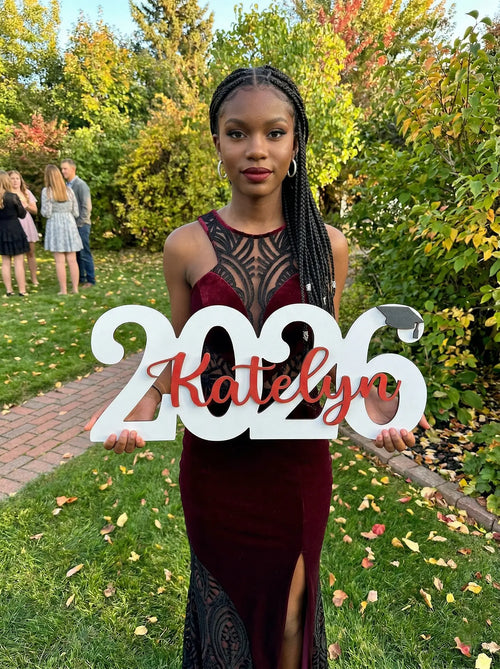 Woman holding a '2026 Katelyn' sign outdoors with greenery in the background