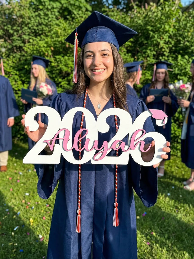 Graduating student holding a '2026 Aliyah' sign outdoors with greenery.