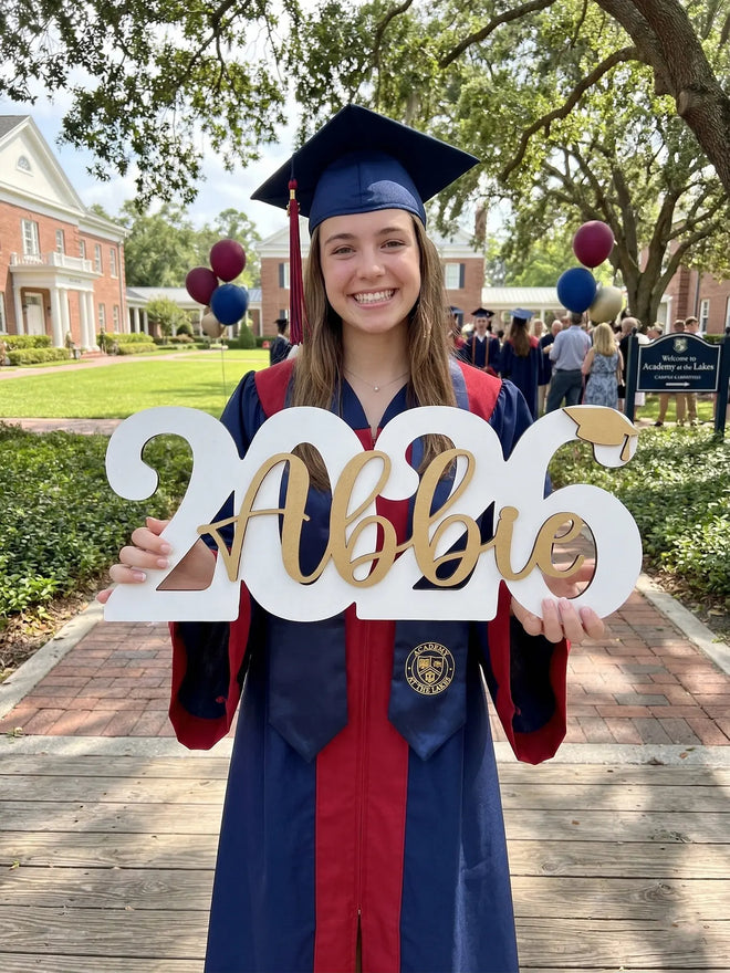Graduate in cap and gown holding '2026 Abby' sign outdoors.