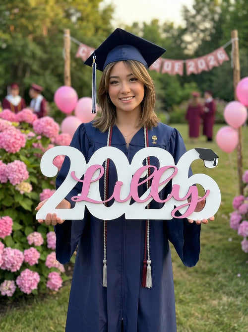 Graduate in cap and gown holding a '2026 Lilly' sign with pink balloons and flowers in the background.