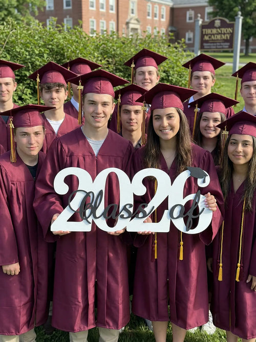 Graduates in maroon caps and gowns holding a '2026 Class of' sign outdoors.