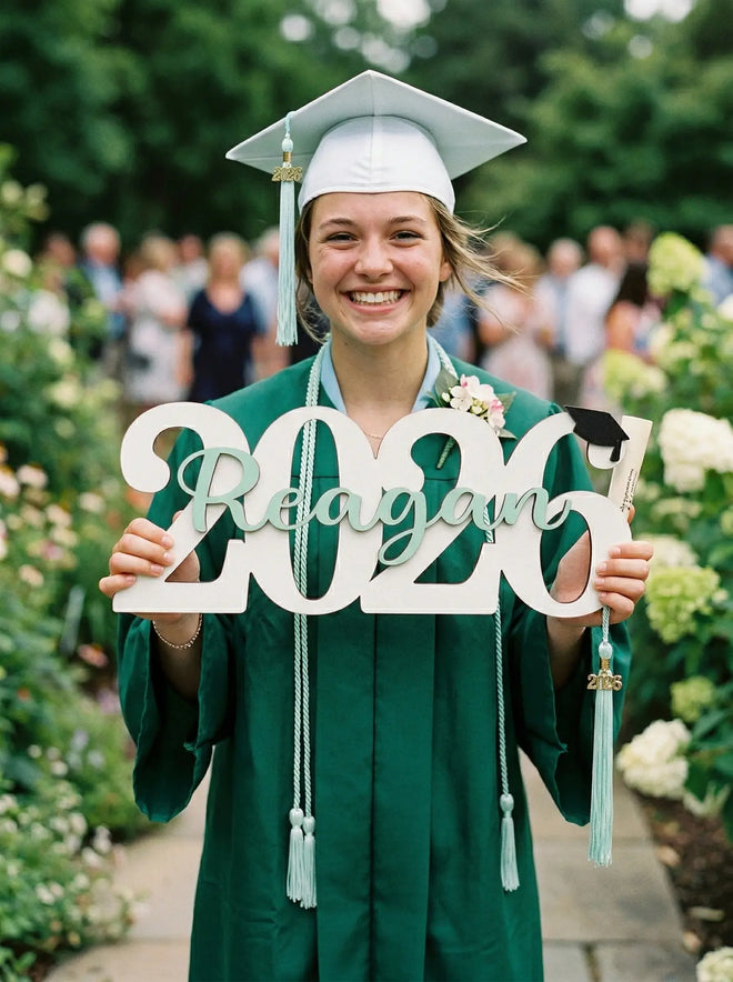 Graduate in cap and gown holding a personalized 2026 graduation year sign. Custom 3D wooden name prop for Class of 2026 senior photo shoots.