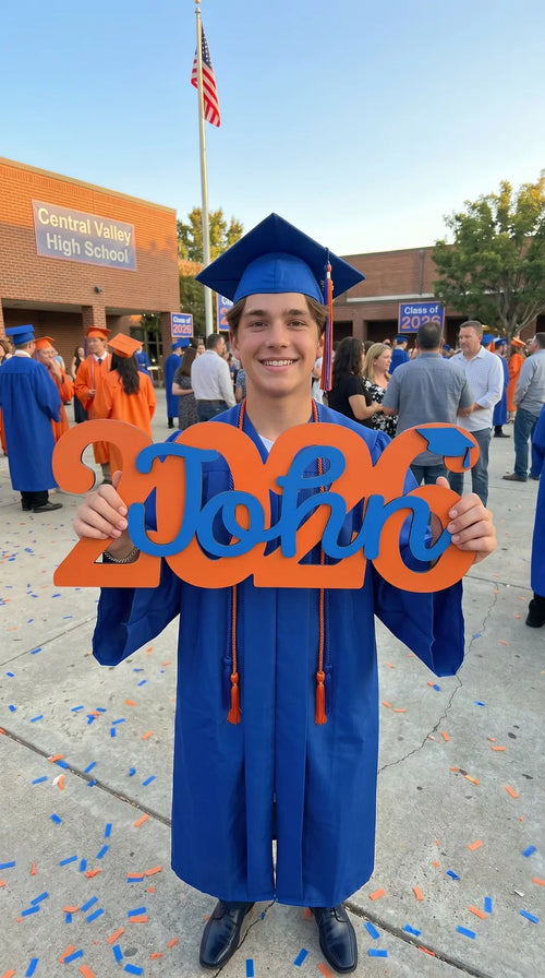 Graduating high school student holding decorative letters spelling 'John 2026' in front of a school building.